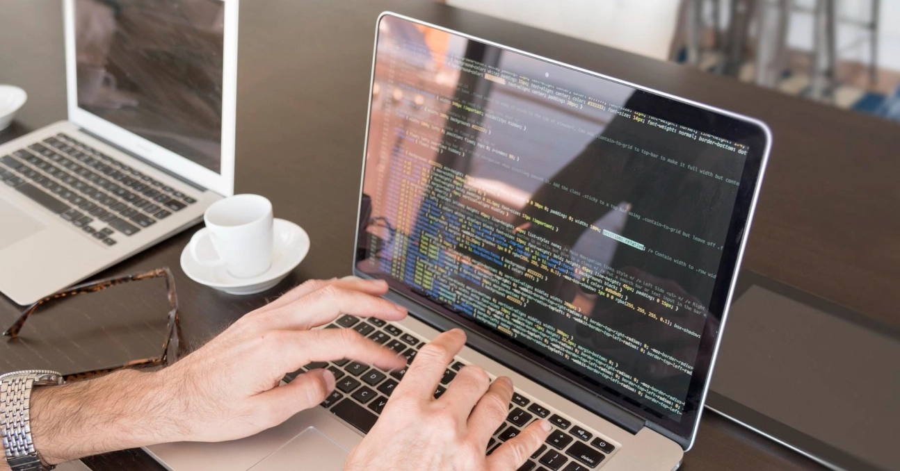 Person typing code on a laptop in a modern workspace with coffee and glasses nearby.