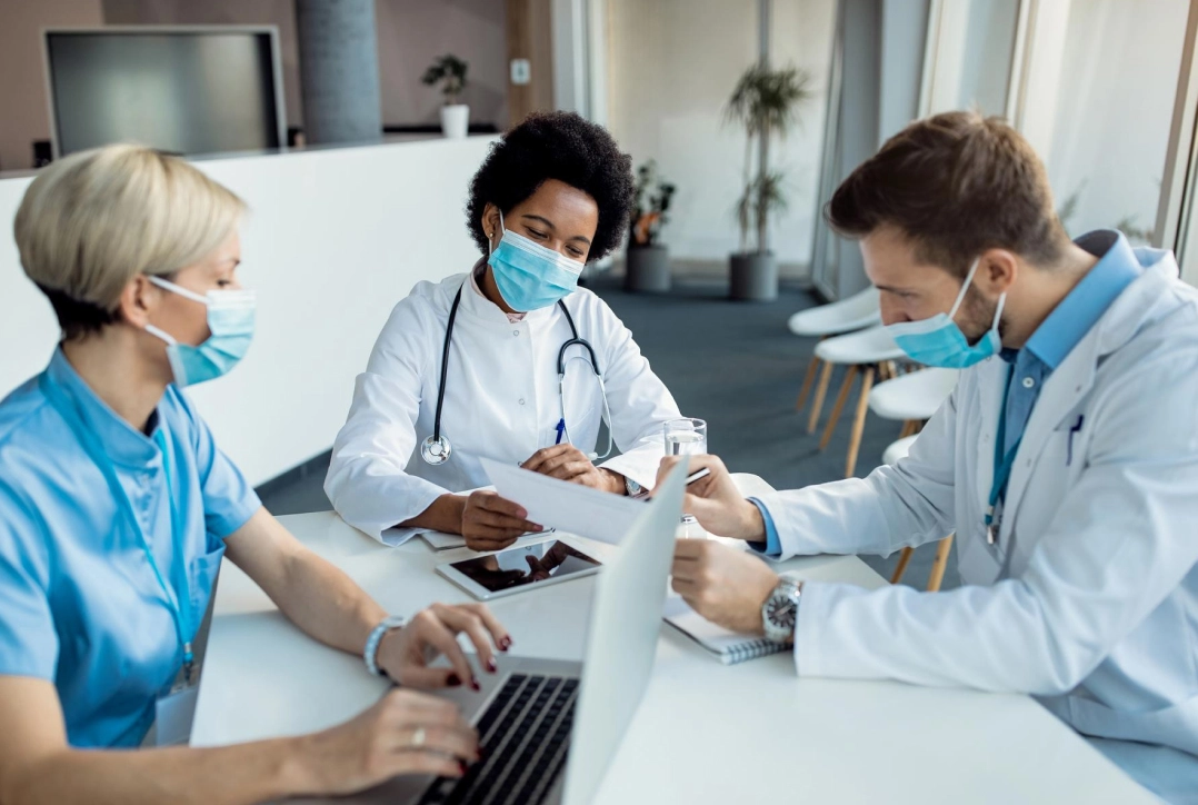 Doctors wearing masks collaborate at a hospital, discussing tasks with a laptop and documents in a bright office space.