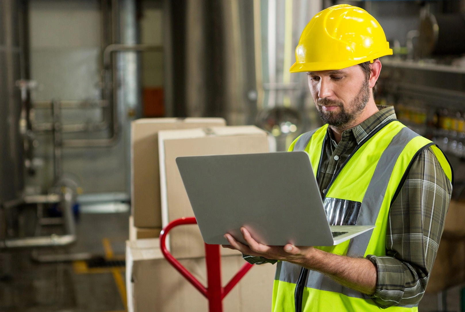 Worker in safety gear using a laptop in a factory setting, surrounded by boxes and industrial equipment.
