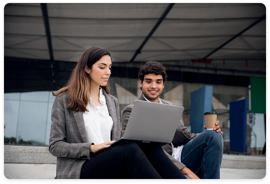 Two professionals sitting on steps, discussing work on a laptop, with a coffee cup, in an outdoor modern setting.