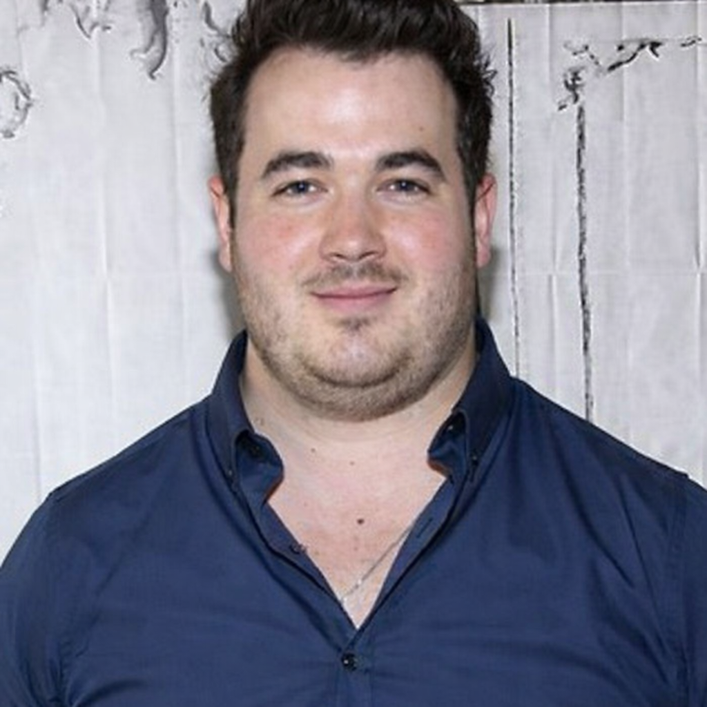 Smiling man in a navy blue shirt standing against a textured wall.
